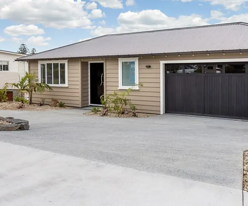 The finished new home in Maraetai from the drive, weatherboard walls, a dark garage door and a new concrete driveway.