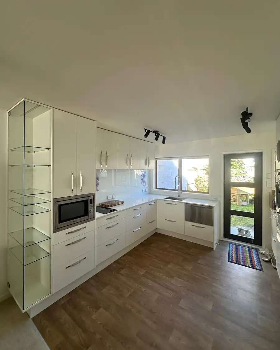 The finished Te Atatū Peninsula kitchen, white cabinets with pull handles, glass display shelves, a built-in microwave, the hob and the sink under the window.